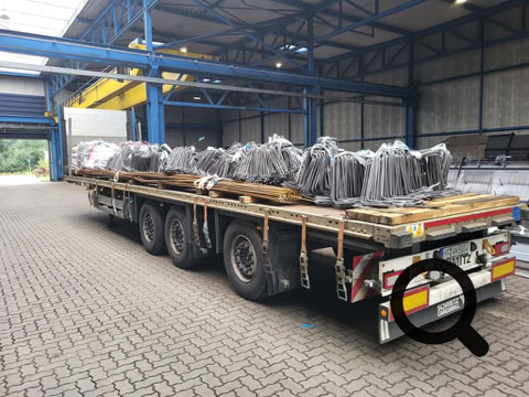 LKW mit offenem Anhänger, beladen mit Metallteilen, in einer Lagerhalle geparkt.
