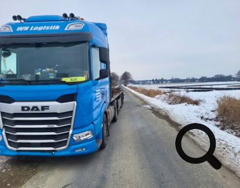 Blauer LKW auf einer schneebedeckten Landstraße, umgeben von Feldern und Bäumen im Hintergrund.