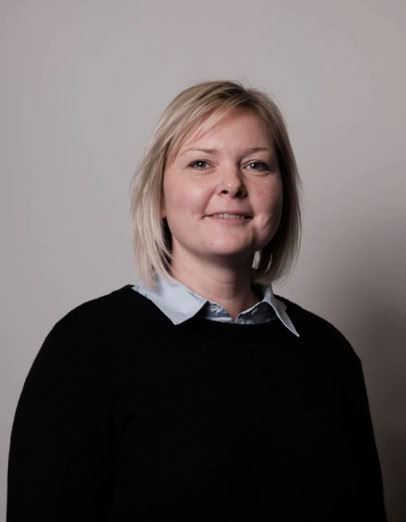 Eine Frau mit blonden Haaren trägt einen schwarzen Pullover über einem hellblauen Hemd vor einem neutralen Hintergrund.