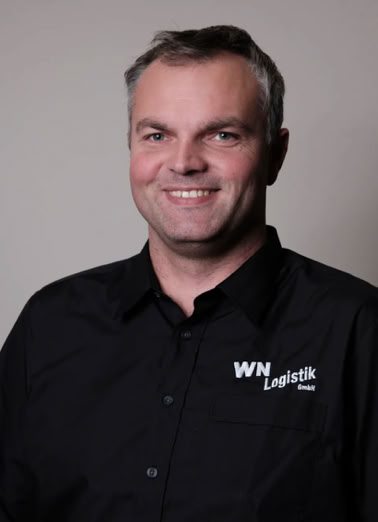 Mann mit grauen Haaren in schwarzem Hemd mit "WN Logistics"-Logo, lächelt vor neutralem Hintergrund.