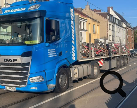 Blauer Lastwagen mit Anhänger auf einer Straße, im Hintergrund Gebäude und blauer Himmel.