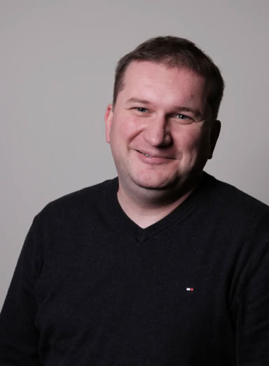 Ein Mann in einem dunklen Pullover lächelt vor einem neutralen Hintergrund.