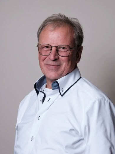 Älterer Mann mit Brille und weißem Hemd vor neutralem Hintergrund.